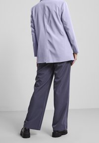 Osoba stojící zády, oblečená v levandulovém blejzru, tmavých širokých kalhotách a černých robustních botách na pozadí s jednobarevným světlým pozadím.