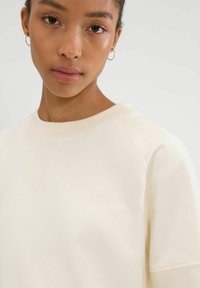 Lichtbeige sweatshirt met geribbelde ronde halslijn, raglanmouwen en een subtiel geperst logo. Zachte stoftextuur met minimale ontwerpdetails.