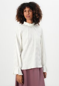 Donna con capelli ricci che indossa una blusa bianca a maniche lunghe con texture e polsini e colletto a volant, abbinata a una gonna malva.