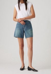 Pantalones cortos de mezclilla en azul claro, de corte relajado con una alta cintura, combinados con una blusa ajustada sin mangas blanca y zapatos planos negros. Textura suave.