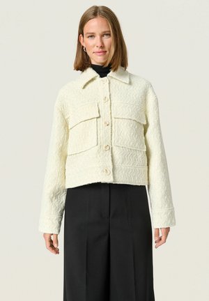 Chaqueta blanca corta de tejido texturizado, con dos bolsillos en el pecho, cierres de botón y un cuello con solapa. Combinada con pantalones negros.