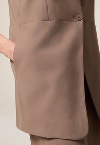 Braun maßgeschneiderte Jacke mit glatter Textur, die eine Seitentasche und ein Knopfdetail in der Nähe der Schulternaht aufweist.