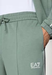 Ανοιχτό πράσινο παντελόνι φόρμας με ελαστική μέση με κορδόνι, πλαϊνές τσέπες και λευκό λογότυπο EA7. Ομα λή υφή με αντιθετικές λευκές διακοσμητικές λεπτομέρειες.