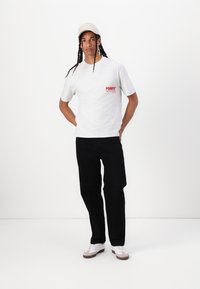 Gray cotton t-shirt with red "POMPEI" logo, black pants, and a beige cap. The model wears white sneakers with brown soles, standing casually.