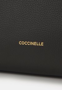 Sort tekstureret læder med en subtil kornstruktur; har guldtekst, der tydeligt staver "COCCINELLE".