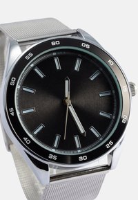 Orologio con quadrante nero, lancette e indici delle ore argentati, cassa rotonda in acciaio inossidabile e cinturino in rete argentata. Presenta una lunetta nera con numeri bianchi.