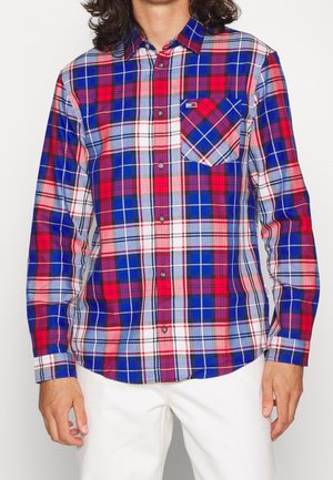 Homme portant une chemise à carreaux à manches longues rouge, bleue et blanche avec une poche poitrine, associée à un pantalon blanc.
