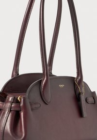 Borsa a mano in pelle color bordeaux con doppi manici, forma arrotondata, texture sottile, dettagli in metallo dorato e un piccolo etichetta con il marchio attaccata.