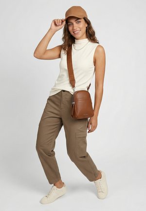 Young woman wearing beige sleeveless top, brown cargo pants, tan cap, white sneakers, and brown crossbody bag, smiling and posing against neutral background.