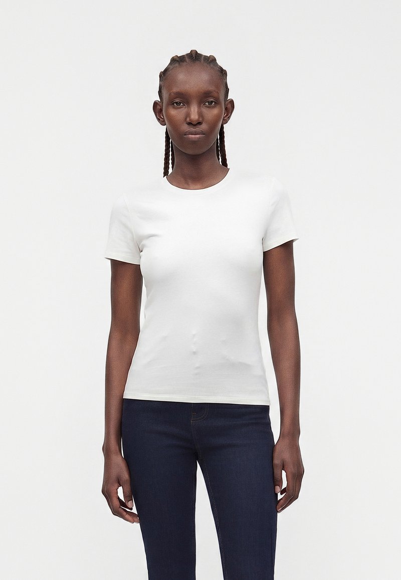Femme aux cheveux tressés portant un t-shirt blanc uni à manches courtes et un jean bleu foncé, debout devant un fond blanc.