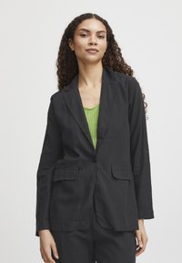 Dunkelgrauer Blazer mit Reverskragen, zwei vorderen Taschen und einer einzigen Knopfverschluss. Trägt man über einem grünen gerippten Oberteil. Glatte Stofftextur.