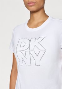 Balta kokvilnas T-kreklu ar lielu, konturētu "DKNY" grafiku sudraba krāsā. Īsās piedurknes un apaļa izgriezne. Gluda tekstūra, brīvs piegriezums.