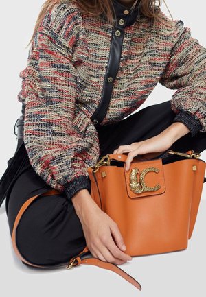 Orangefarbene Leder-Bucket-Bag mit strukturiertem, goldfarbenem Metallakzent und schwarzen Details. Modell trägt ein gemustertes Strickoberteil mit aufgepufften Ärmeln.