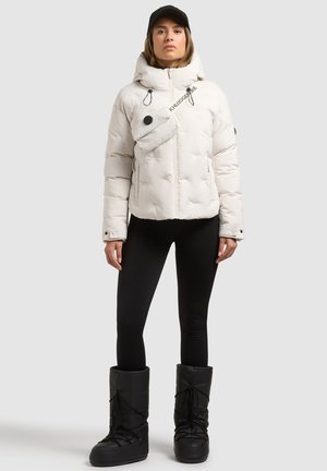 Femme portant une veste matelassée blanche avec capuche, des leggings noirs, des bottes d'hiver noires et une casquette noire, debout devant un fond uni.