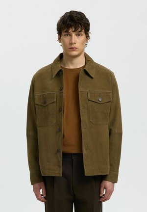 Jeune homme aux cheveux foncés portant une veste boutonnée en daim marron sur une chemise marron et un pantalon foncé, debout devant un fond uni.