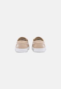 Beige Leinensneaker mit weißen Gummisohlen, zwei hinteren Ösen und einem Logo an der Ferse. Glatte Textur und niedriges Profil.