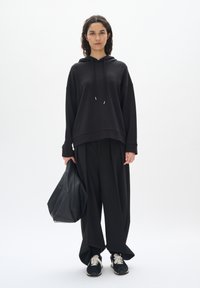 Felpa nera oversize con cappuccio e lacci, abbinata a pantaloni neri larghi e sneakers nere. Il modello porta una borsa di pelle nera.