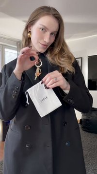 Llavero dorado con diseño de cadena y un pequeño detalle naranja, sostenido por una persona con un abrigo negro, que también muestra una bolsa blanca.