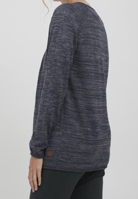 Tummansininen pitkähihainen villapaita, jossa on teksturoitu, marmorikuvioitu neulominen. Siinä on pyöreä pääntie ja pieni logopatchi helman lähellä.