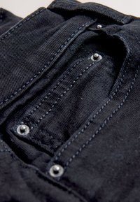 Gros plan sur un tissu denim noir montrant une petite poche avec des rivets métalliques et des détails de couture visibles.