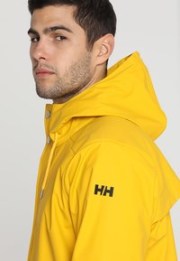 Geel waterdichte jas met een capuchon, met een gladde textuur, drukknopen en een zwart logo op de linker schouder.