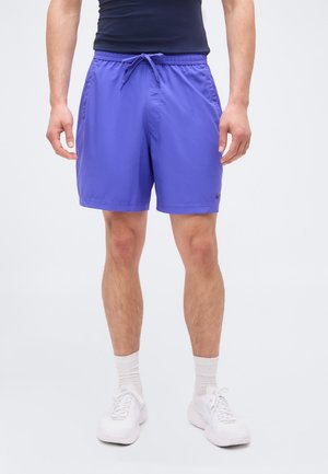 Άνδρας που φοράει μωβ αθλητικά σορτς με κορδόνι, λευκές κάλτσες, λευκά αθλητικά παπούτσια και σκούρο αμάνικο τοπ, στέκεται μπροστά σε απλό φόντο.