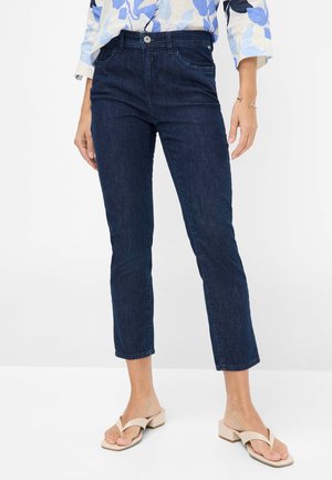 Woman wearing dark blue cropped jeans and beige heeled sandals, standing against a plain white background.