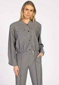 Een blonde vrouw draagt een grijze cropped jas met knopen en elastische taille, gecombineerd met bijpassende hoog getailleerde plooibroek, met één hand in haar zak.