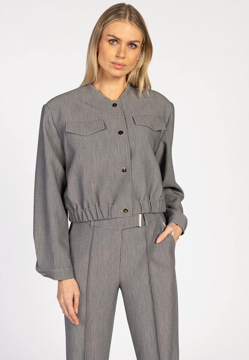 Een blonde vrouw draagt een grijze cropped jas met knopen en elastische taille, gecombineerd met bijpassende hoog getailleerde plooibroek, met één hand in haar zak.