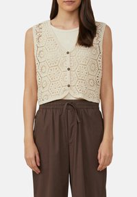 Häkelweste in Creme mit hexagonalen Mustern, mit V-Ausschnitt und Knopfverschluss. Über einem hellen Top getragen und mit braunen Hosen kombiniert.