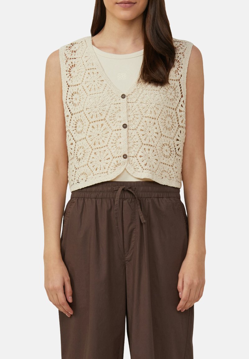Häkelweste in Creme mit hexagonalen Mustern, mit V-Ausschnitt und Knopfverschluss. Über einem hellen Top getragen und mit braunen Hosen kombiniert.