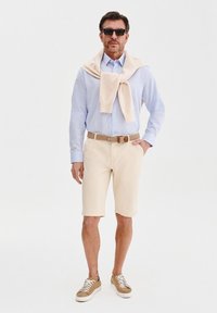 Beżowe szorty w połączeniu z jasnoniebieską koszulą z guzikami. Na ramionach zarzucony jest kremowy sweter. Akcesoria to okulary przeciwsłoneczne i brązowy pasek.