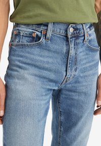 Ljusblå denimjeans med en lätt blekning, femficksdesign och orange sömmar som accent. Har knappar och dragkedjeflyclosures.