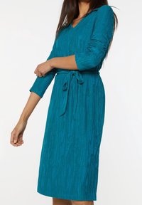 Teal stickad klänning med V-ringning, trekvartsärmar, bälte i midjan, och texturerade vertikala ränder.