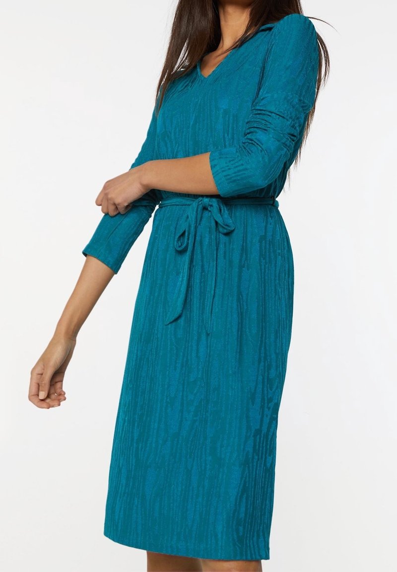 Teal stickad klänning med V-ringning, trekvartsärmar, bälte i midjan, och texturerade vertikala ränder.