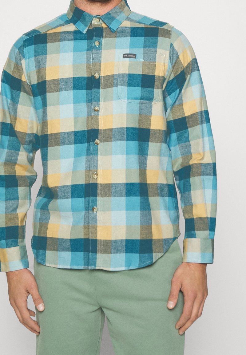Homme portant une chemise à manches longues à carreaux bleu, sarcelle, beige et jaune, et un pantalon vert clair.