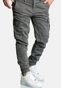 Mężczyzna w szarych dżoggerach z elastycznymi mankietami i białych butach Nike Air Force 1, stojący z rękami w kieszeniach.