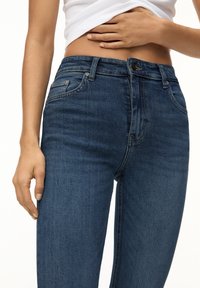Jeans de mezclilla azul oscuro con corte slim, que cuentan con un cierre de botón, pasadores para cinturón y una textura suave. Manos descansando sobre la cinturilla.
