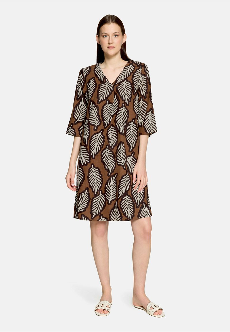 Femme debout portant une robe marron mi-longue avec de grands motifs de feuilles blanches et des sandales blanches ouvertes devant un fond uni.