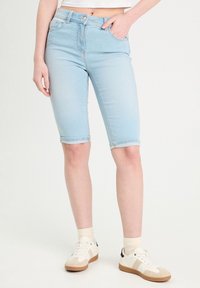 Bermudas en denim bleu clair au design ajusté, avec cinq poches et un ourlet roulé, accompagnées de baskets blanches et de chaussettes crème.