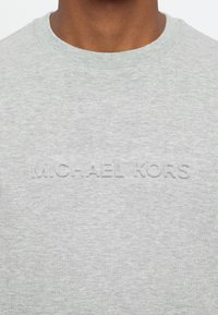 Sudadera gris con cuello redondo, que presenta un logo en relieve de "MICHAEL KORS" en el frente. Confeccionada en una suave tela texturizada.