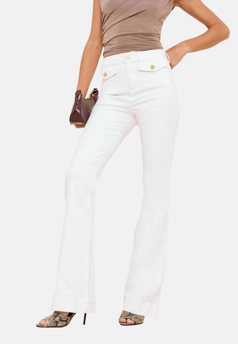 Kvinde iført højtaljede hvide viddebukser med gyldne knapper, taupe ærmeløs top, højhælede sko med slangeskindsmønster og holder en brun håndtaske.