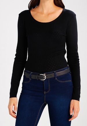 Femme portant un haut noir moulant à manches longues rentré dans un jean bleu foncé avec une ceinture noire à boucle argentée.