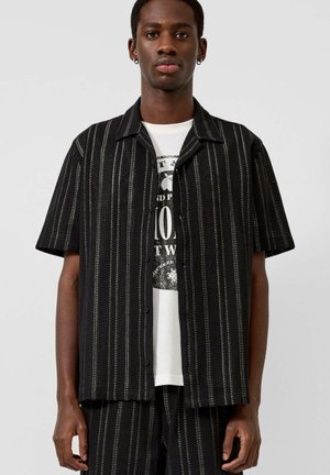 Młody mężczyzna ubrany w czarną koszulę z krótkim rękawem, z białymi pionowymi paskami, na białej koszulce z nadrukiem na jednolitym tle.