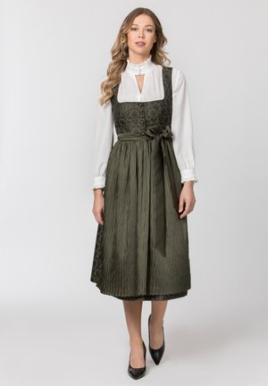 Frau trägt eine weiße Bluse unter einem dunkelgrünen, geblümten Dirndl-Kleid mit einer plissierten Schürze, die in der Taille gebunden ist, und schwarzen High Heels.