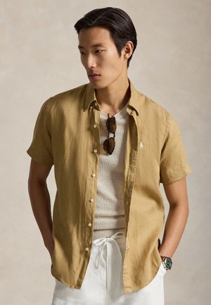 Νέος άνδρας που φοράει μια καμηλό κοντομάνικη πουκάμισο με κουμπιά πάνω από ένα μπεζ πλεκτό τοπ, λευκά παντελόνια με κορδόνι, γυαλιά ηλίου κρεμασμένα στο πουκάμισο και ένα ρολόι χειρός.