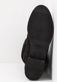 Czarna botka spoczywająca na białej powierzchni i ścianie, z widoczną żebrowaną podeszwą i nadrukowanym w środku rozmiarem 37.