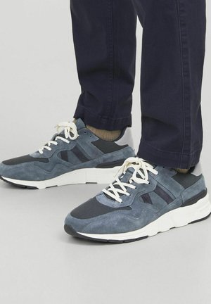 Grå ruskinds sneakers med mesh-paneler, hvid gummisål og kontrasterende mørkegrå detaljer. Har hvide snørebånd og en polstret tunge.