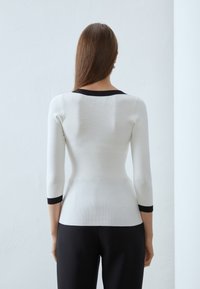 Maglione bianco con maniche a tre quarti, con scollatura rotonda nera e polsini neri. Tessuto liscio con una silhouette aderente.