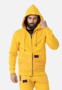 Gelbe Zip-Hoodie mit einer Kapuze mit Kordelzug, Fronttaschen und kontrastierendem, strukturiertem Einsatz. Mit schwarzem Reißverschluss und Branding-Akzenten.